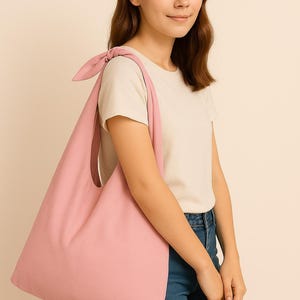 May include: A light pink tote bag with a knotted shoulder strap. The bag is made of a soft fabric and has a simple, minimalist design. The bag is being carried by a person wearing a white t-shirt and blue jeans. The logo "KM with love" is in the upper left corner.