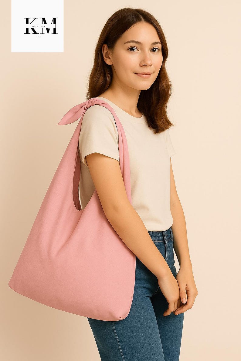 May include: A light pink tote bag with a knotted shoulder strap. The bag is made of a soft fabric and has a simple, minimalist design. The bag is being carried by a person wearing a white t-shirt and blue jeans. The logo "KM with love" is in the upper left corner.