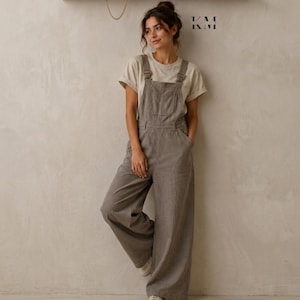 May include: A woman wearing a light brown t-shirt and gray overalls with wide legs. The overalls have adjustable straps and pockets. The woman is leaning against a light-colored wall. A wooden shelf with jewelry and small objects is above her head.