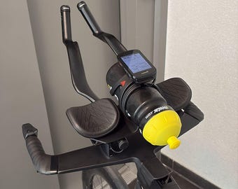 Puede incluir: Un cockpit de bicicleta de triatlón negro con manillar aerodinámico, apoyabrazos negros, una botella de agua negra con tapón amarillo brillante etiquetada como Isostar.com y un ciclocomputador digital montado en el centro.