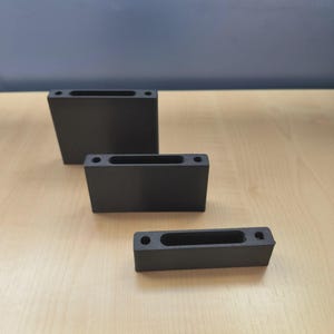 May include: Three black rectangular blocks of varying sizes are arranged on a light wood surface. Each block has a long, narrow, rectangular opening and two small circular holes. The background is a solid blue-grey.