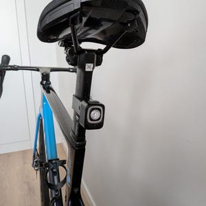May include: A black bicycle seat mounted on a black seat post with a square rear light attached. The bicycle frame is black and blue, featuring a bottle cage and a small QR code sticker on the seat post. The bike is positioned upright.