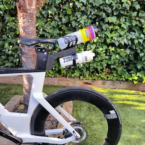 May include: A white and black road bike with two water bottles attached. One bottle is white with black text, the other is clear with a colorful design. The bike has a black wheel and a black seat.