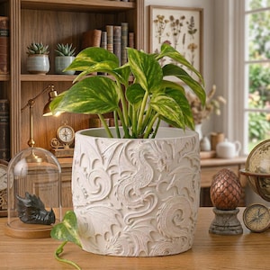 May include: A cream-coloured ceramic planter, adorned with a dragon design, containing a green plant with yellow markings. The decorative planter is a stylish addition to any home interior.