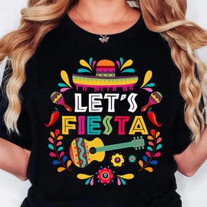 May include: Black t-shirt with a colorful "Let's Fiesta" design. The graphic includes a sombrero, maracas, chili peppers, a guitar, and floral elements. The design is vibrant with yellow, red, green, and blue.