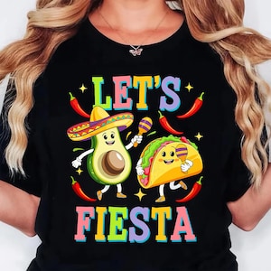 May include: Black t-shirt with a colorful graphic that says "LET'S FIESTA". The design features a smiling avocado wearing a sombrero and a taco holding maracas, with chili peppers and stars.