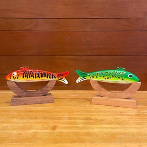 May include: Two wooden fish figurines on display stands. The fish are painted in bright colors. One fish is orange and black with a red fin. The other fish is green with black spots.