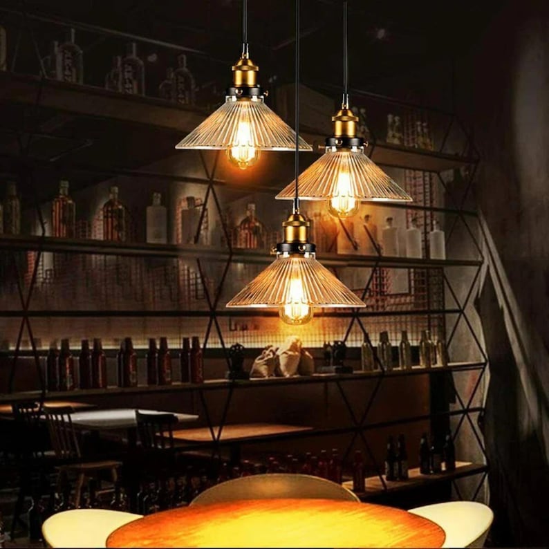 May include: Three glass pendant lights with a vintage industrial design. The lights have a clear glass shade with a ribbed texture and a black metal frame. The lights are hanging from black cords and are illuminated with warm white light bulbs.