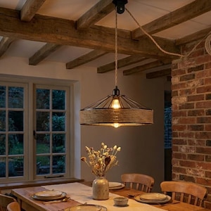 May include: A vintage-style pendant light with a metal frame and a rope-wrapped section. The light hangs from a rope cord above a dining table. The room features exposed wooden beams, a brick wall, and a vase of dried flowers. The light is on.