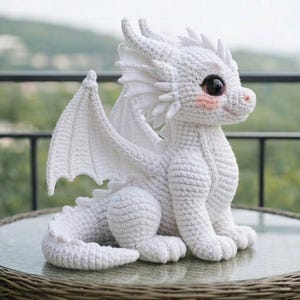 May include: A white crocheted dragon figurine with large, dark eyes and pink cheeks. The dragon has detailed wings and sits on a round, glass-topped table. The dragon is a handmade item.