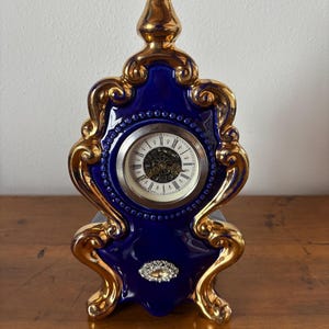 Pode incluir: Um relógio decorativo de porcelana azul e dourada. O mostrador redondo exibe algarismos romanos. O relógio tem detalhes dourados elaborados e uma pequena joia na base. Mede aproximadamente 15 cm de altura.