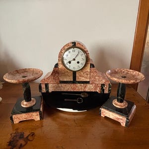 Puede incluir: Un reloj antiguo con candelabros a juego. El reloj tiene una esfera redonda con números romanos y un patrón similar al mármol en tonos rojos, negros y crema. Los candelabros tienen un diseño similar y están sobre bases negras.