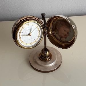 Könnte beinhalten: Eine Vintage-Uhr in Goldtönen mit weißem Zifferblatt und schwarzen Ziffern. Die Uhr ist aufklappbar und öffnet sich, um ein kleines Foto zu zeigen. Die Uhr steht auf einem silber-goldenen Sockel.