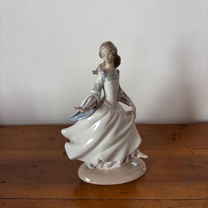 Può includere: Statuina in porcellana di una donna in un abito bianco fluente con accenti blu e rosa. La statuina è in posa in una danza aggraziata, con le braccia tese e la gonna che turbina. La base è di colore beige chiaro.
