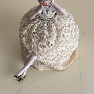 Può includere: Una bambola vintage in porcellana con parrucca grigia e lineamenti delicati. La bambola indossa un abito di pizzo color crema, calze bianche e scarpe dorate. La bambola è seduta con le braccia appoggiate sull'abito.