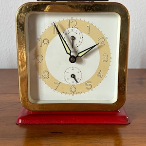 May include: A vintage gold-toned alarm clock with a square face and a red base. The clock face is white with a cream-colored ring, black hands, and black numerals. The clock reads "Made in Germany".