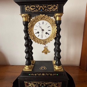 Op de afbeelding: Een antieke zwarte mantelklok met gouden accenten. De klok heeft een ronde wijzerplaat met zwarte Romeinse cijfers en sierlijke gouden details. De klok heeft een decoratieve slinger en een sleutel.