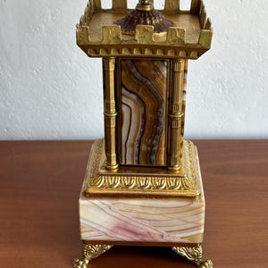 May include: An antique decorative box with a castle-like design. The box has a rectangular base with ornate gold-coloured feet and trim. The central column is made of layered brown, white, and tan stone, topped with a gold-coloured, castle-like structure.