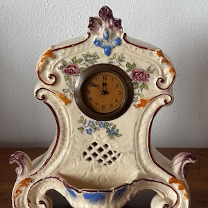 Puede incluir: Reloj de chimenea de cerámica color crema con detalles florales y de volutas. La esfera del reloj es redonda con números y manecillas negras. El reloj tiene un pequeño estante en la base y está adornado con detalles rosas, azules y naranjas.