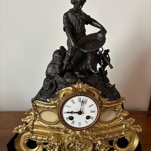 Könnte beinhalten: Antike bronzefarbene Kaminuhr mit einer Figur, die eine Pfanne hält, auf einem dekorativen Sockel mit einem Zifferblatt. Die Uhr hat einen goldfarbenen Rahmen mit aufwendigen Details und ein weißes Zifferblatt mit schwarzen römischen Ziffern.