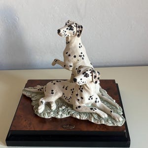 Könnte beinhalten: Dekorative Figur mit zwei Dalmatinerhunden. Einer steht mit erhobener Pfote, der andere liegt. Die Hunde sind weiß mit schwarzen Flecken, auf einem Holzsockel mit schwarzem Rand.