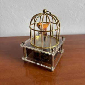 Puede incluir: Una caja de música decorativa vintage con un pequeño pájaro naranja dentro de una jaula dorada. La jaula se asienta sobre una base de acrílico transparente con detalles dorados. El pájaro está posado en una pequeña plataforma.