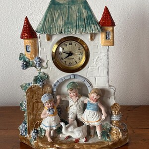Puede incluir: Un reloj decorativo de cerámica con una torre blanca con tejado verde y torretas rojas. La esfera del reloj es dorada. Tres figuras de niños están delante del reloj, con un ganso. El reloj está sobre una superficie de madera.