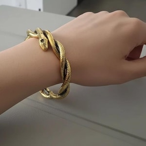 Puede incluir: Un brazalete dorado con forma de serpiente enrollada alrededor de la muñeca. La cabeza de la serpiente es visible, con escamas y ojos detallados. El brazalete tiene una superficie texturizada y un diseño retorcido.