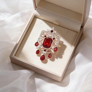 May include: A red gemstone pendant with a gold-coloured frame and clear accent stones, displayed in a beige jewellery box. The pendant features a large, central red stone and smaller, teardrop-shaped red stones hanging below.