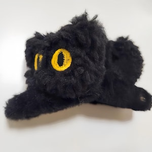 Puede incluir: Juguete de gato de peluche negro con ojos amarillos. El gato está acostado de lado con la cabeza girada hacia la derecha.