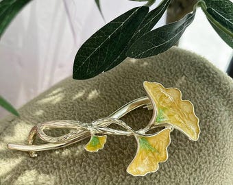 May include: A silver-toned brooch featuring a stylized ginkgo leaf design. The leaves are a vibrant yellow with green accents, set against a silver metal stem. The brooch is a decorative accessory.