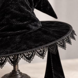 May include: A black velvet witch hat featuring a pointed crown and a wide brim. The edge of the brim is finished with a detailed black lace trim that hangs down in a scalloped pattern. The fabric has a soft, crushed texture throughout the surface.
