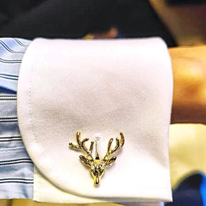 Può includere: Gemelli dorati a forma di testa di cervo con dettagli in cristallo trasparente, su un polsino di camicia bianca. La manica della camicia presenta un motivo a righe blu chiaro. Un tocco di eleganza per l'abbigliamento formale.