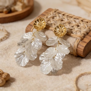 May include: A pair of elegant earrings featuring a gold-colored floral stud and cascading white, petal-shaped dangles. The earrings are set against a neutral background, highlighting their delicate design and intricate details. These earrings are perfect for adding a touch of sophistication.