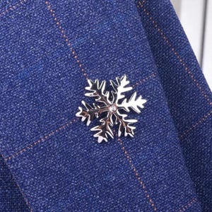 May include: A silver snowflake brooch with a small crystal center is pinned to a blue plaid jacket. The brooch has intricate details and a polished finish, adding a touch of winter elegance to the garment.
