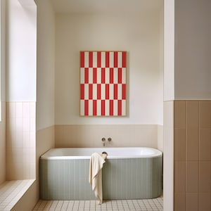 May include: A bathroom scene featuring a rectangular framed artwork with a red and white striped pattern. Below, a light green tiled bathtub with a white interior and a beige towel hanging over the edge. The walls are a light beige color.