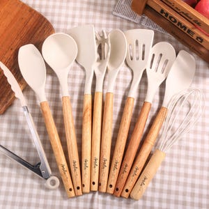 Könnte beinhalten: Eine Sammlung von Küchenutensilien mit cremefarbenen Silikonköpfen und Griffen aus Naturholz. Das Set umfasst einen Spatel, einen Löffel, einen Schneebesen und eine Zange, angeordnet auf einer karierten Oberfläche. Die Griffe tragen den Aufdruck "Amelia".