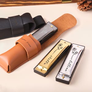 Puede incluir: Armónicas en estuches de cuero. Se ven dos armónicas, una dorada y otra plateada, cada una con un diseño floral y un nombre. Los estuches son marrones y negros, uno de ellos con la inscripción "Together forever".