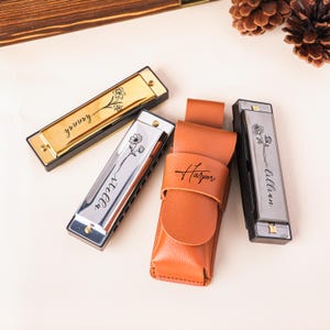 Puede incluir: Cuatro armónicas con nombres personalizados y diseños florales. Dos armónicas tienen cubiertas doradas y dos plateadas. También hay un estuche de cuero marrón con el nombre "Harper".