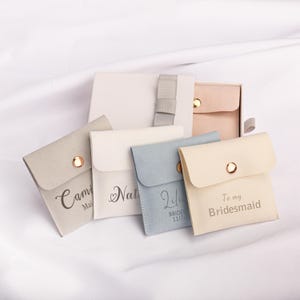 Puede incluir: Cinco pequeñas cajas de regalo cuadradas de tela con botones dorados. Las cajas son de varios colores, incluyendo gris, blanco, azul y beige. Cada caja tiene un mensaje personalizado en una fuente cursiva. Los mensajes incluyen "Camille Maid of Honor", "Natalie", "Lili Bride 11/17", y "To my Bridesmaid".