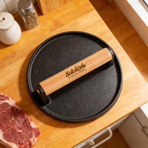 Puede incluir: Prensa para parrilla redonda de hierro fundido negro con un mango de madera marrón claro. El mango tiene el texto "PARIS BBQ" y un diseño de llama. La prensa está sobre una superficie de madera.