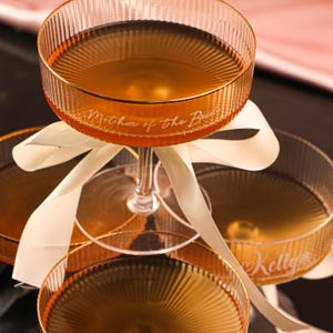 Puede incluir: Primer plano de tres copas de champán llenas de un líquido dorado. Las copas tienen un diseño acanalado y bordes dorados. La copa superior tiene el texto "Mother of the Bride" grabado, mientras que la copa inferior tiene el nombre "Kelly". Una cinta color crema está atada alrededor de los tallos.