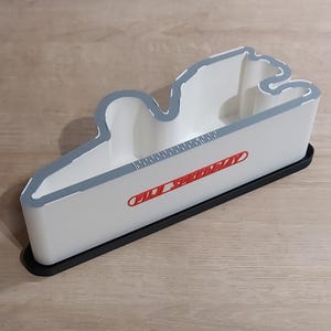 Könnte beinhalten: Ein weißer, wolkenförmiger Behälter mit grauem Umriss und einem rot-weißen Logo. Der Behälter steht auf einer schwarzen rechteckigen Basis. Der Innenraum ist weiß, und der Behälter steht auf einer hellbraunen Holzoberfläche.