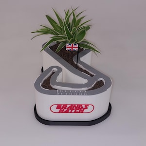 May include: White planter shaped like the Brands Hatch race track with a small green plant growing inside. The planter has a black base and the words "Brands Hatch" are printed in red on the front.
