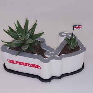 Puede incluir: Macetero blanco con forma de circuito de Silverstone con dos pequeñas suculentas y una pequeña bandera británica.