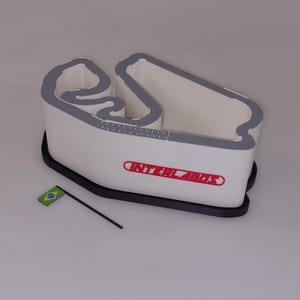 以下が含まれることがあります： 「INTERLAGOS」の赤い文字が入った、白とグレーのインテルラゴスレーストラックの模型。小さなブラジル国旗が模型の近くに配置されています。トラックは黒いベースと灰色の縁取りがあります。