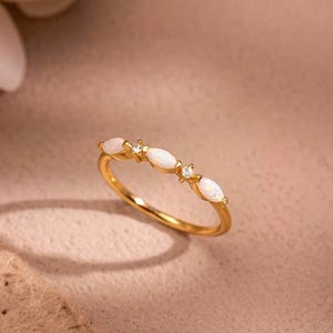 Könnte beinhalten: Ein goldener Ring mit filigranem Design. Der Ring zeigt eine Reihe von oval geformten weißen Edelsteinen, die mit kleineren, klaren Steinen durchsetzt sind. Der Ring ist vor einem weichen, neutralen Hintergrund platziert, mit einem Hauch einer Blume in der oberen linken Ecke.