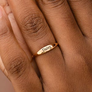 Può includere: Un anello dorato con il nome "Daisy" inciso. L'anello, di forma ovale, è indossato su un dito. L'immagine è un primo piano di una mano che mostra l'anello. L'anello è d'oro.