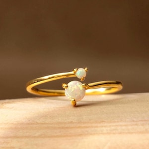 May include: A gold ring featuring two white opal gemstones. The ring has a slender, open design, with one opal at the centre and a smaller one above. The ring is presented on a wooden surface.