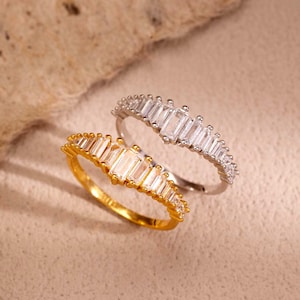 Peut inclure: Deux bagues, une dorée et une argentée, sont présentées. Les deux bagues comportent une rangée de pierres précieuses rectangulaires et transparentes. La bague dorée a une teinte jaune chaude, tandis que la bague argentée a une finition métallique brillante.